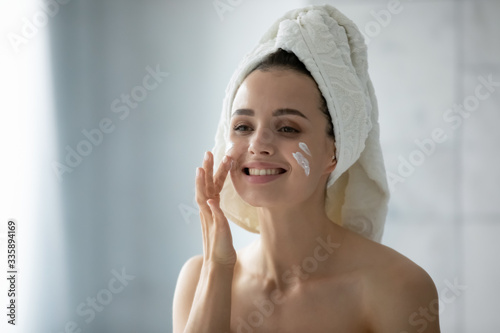 Happy millennial girl in towel after shower look in mirror apply moisturizing facial cream, smiling young woman do daily morning beauty routine procedures or treatment, skincare concept