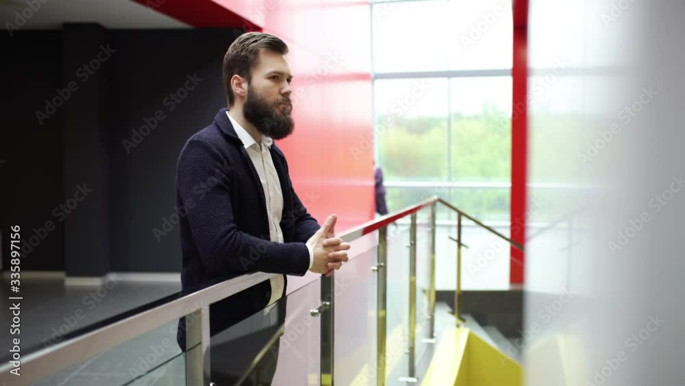 Thoughtful bearded man is standing with clasped together hands near glass fence in modern building. Close-up side view of pensive bearded guy leaning on fence inside business center. Thoughtful bearded man is standing with clasped together hands near glass fence in modern building. Close-up side view of pensive bearded guy leaning on fence inside business center.