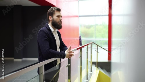 Thoughtful bearded man is standing with clasped together hands near glass fence in modern building. Close-up side view of pensive bearded guy leaning on fence inside business center.