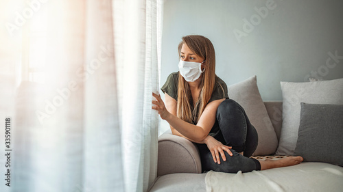 Sad lonely girl isolated stay at home in protective sterile medical mask on face looking at window, bored woman because of Chinese pandemic coronavirus virus covid-19. Quarantine, prevent infection photo