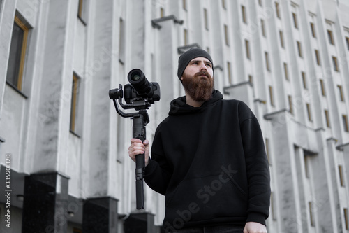 Bearded Professional videographer in black hoodie holding professional camera on 3-axis gimbal stabilizer. Filmmaker making a great video with a professional cinema camera. Cinematographer. photo