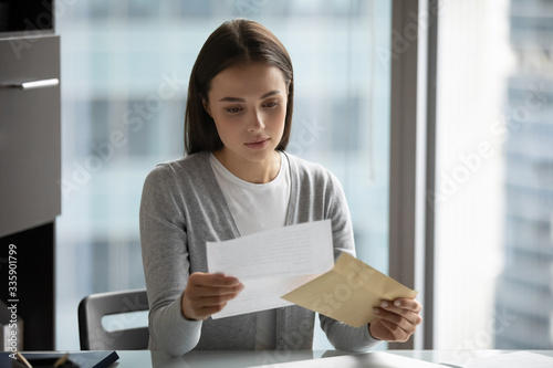 Pensive young woman sit at desk in office open envelope read post correspondence, focused thoughtful millennial female get postal letter consider offer or suggestion, promotion or dismissal notice