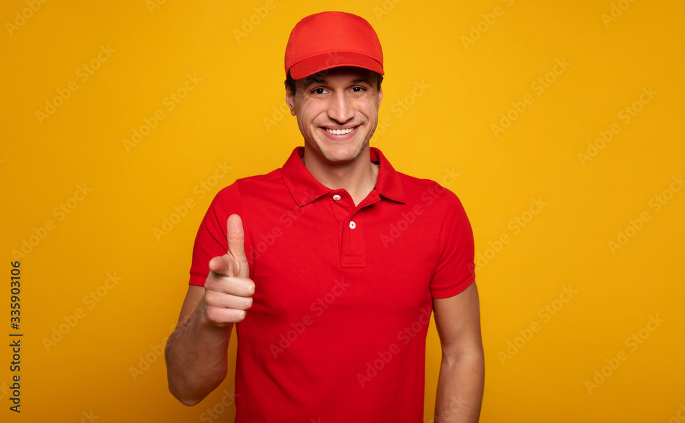 Handsome young happy courier or delivery man in red uniform is posing isolated on yellow background.