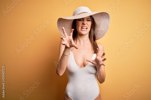 Young beautiful brunette woman on vacation wearing swimsuit and summer hat smiling funny doing claw gesture as cat, aggressive and sexy expression
