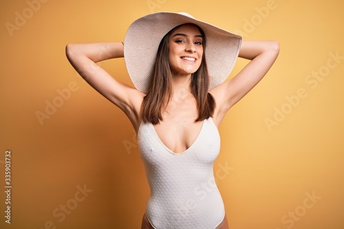 Young beautiful brunette woman on vacation wearing swimsuit and summer hat relaxing and stretching, arms and hands behind head and neck smiling happy