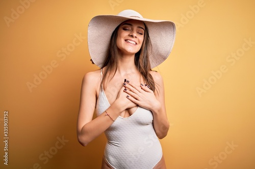 Young beautiful brunette woman on vacation wearing swimsuit and summer hat smiling with hands on chest with closed eyes and grateful gesture on face. Health concept.