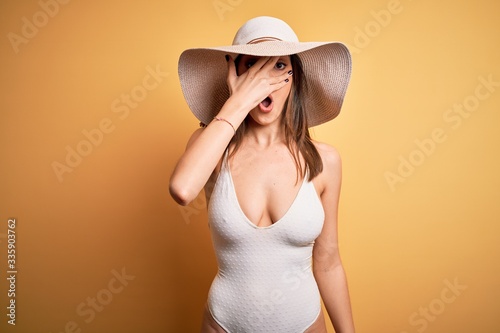 Young beautiful brunette woman on vacation wearing swimsuit and summer hat peeking in shock covering face and eyes with hand, looking through fingers with embarrassed expression.
