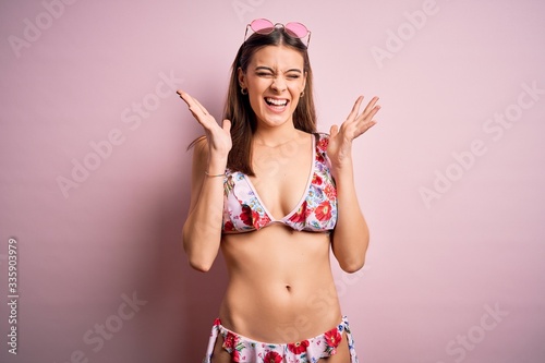 Young beautiful woman on vacation wearing bikini and sunglasses over pink background celebrating mad and crazy for success with arms raised and closed eyes screaming excited. Winner concept
