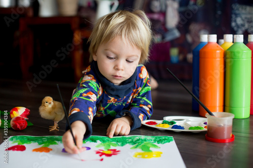Sweet toddler blond child, boy, painting with colors, little chicks walking around him, making funny prints