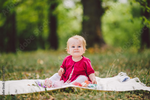 Little baby girl Caucasian ethnicity blond one year old from birth sits on a plaid on green grass in the park. Child picnic in the autumn forest. Beautiful cute joyful funny baby sitting on blanket © Elizaveta