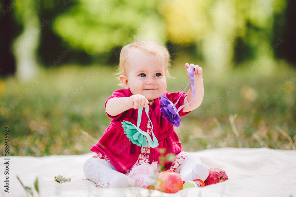 Beautiful Girl 1 Years Old Sitting On The Plaid In The Park. Child outdoor. Baby at picnic in forest. Portrait of a beautiful baby girl sitting on green meadow. Pretty little girl