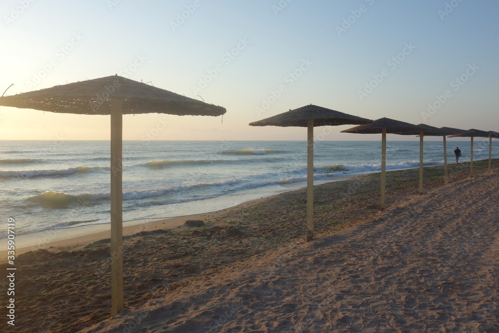 Row of reed parasols along sea shore and single man walking, in Vama Veche, Romania, early in the morning