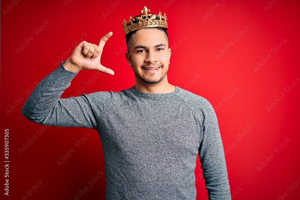 Young handsome man wearing golden crown of prince over isolated red background smiling and confident gesturing with hand doing small size sign with fingers looking and the camera. Measure concept. Young handsome man wearing golden crown of prince over isolated red background smiling and confident gesturing with hand doing small size sign with fingers looking and the camera. Measure concept.