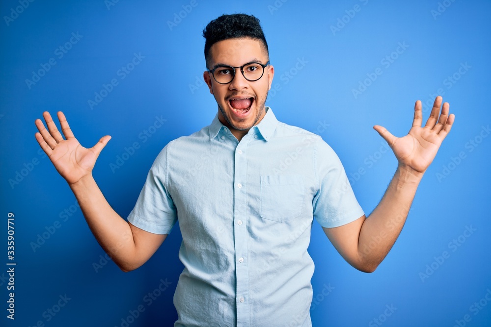 Fototapeta premium Young handsome man wearing casual summer shirt and glasses over isolated blue background celebrating crazy and amazed for success with arms raised and open eyes screaming excited. Winner concept