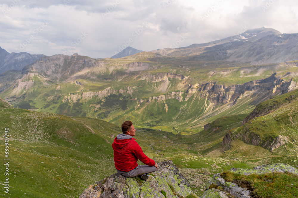 Young man in a red jacket on the stones looks at a magnificent view of the Alps mountains.