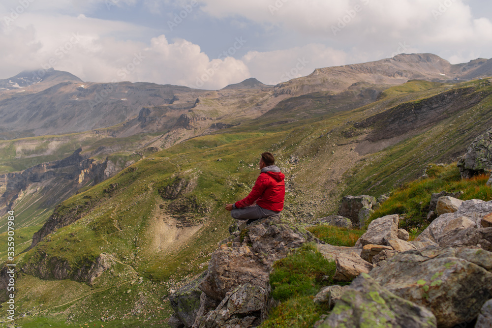 Young man in a red jacket on the stones looks at a magnificent view of the Alps mountains. Young man in a red jacket on the stones looks at a magnificent view of the Alps mountains.