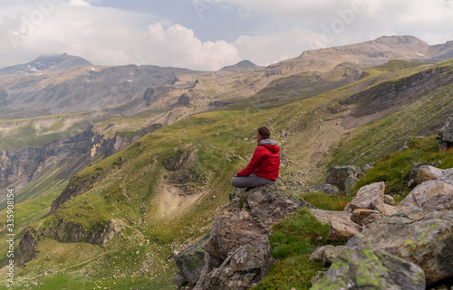 Young man in a red jacket on the stones looks at a magnificent view of the Alps mountains.