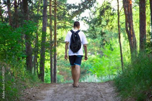 A guy in a white T-shirt, dark shorts and with a backpack walks a sand trail in the forest on a sunny summer day.