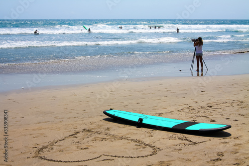 A surfboard is lying on the beach. Next to it, a large heart is drawn in the sand. Good weather, small waves. People learn to surf in the ocean.