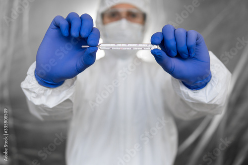 Doctor in white protective suit, medical mask and rubber gloves is holding a thermometer in coronavirus pandemic. Epidemic, pandemic of coronavirus covid 19. Measuring and indicating temperature.