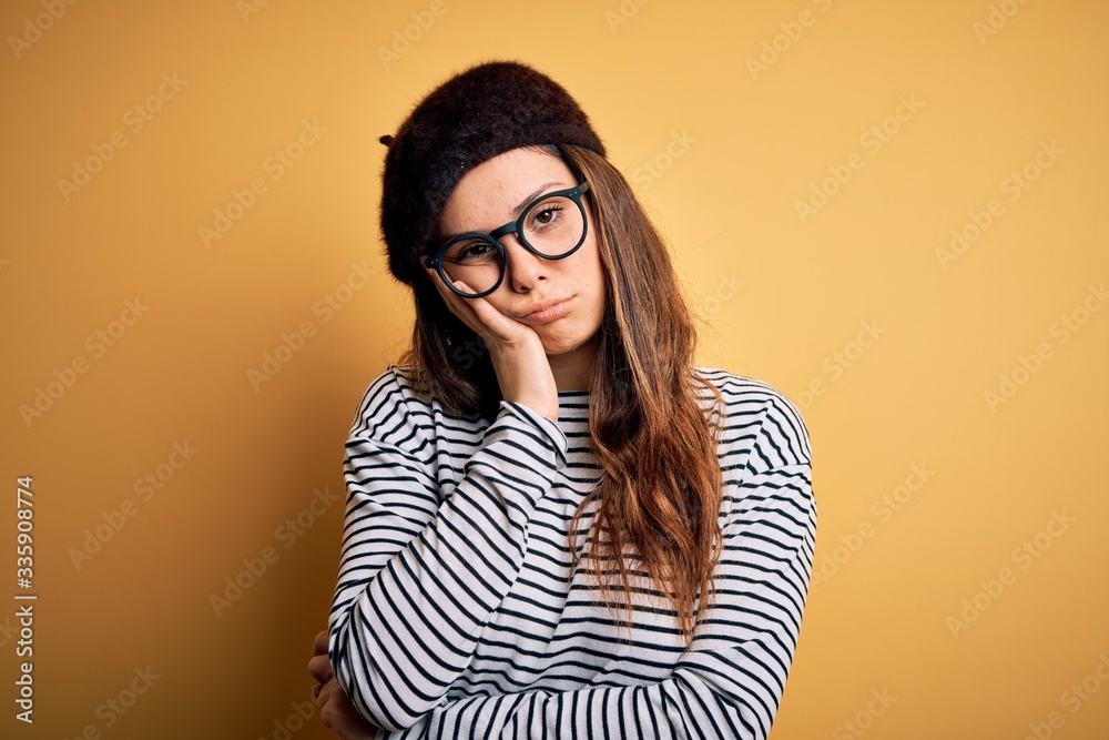 Young beautiful brunette woman wearing french beret and glasses over yellow background thinking looking tired and bored with depression problems with crossed arms.
