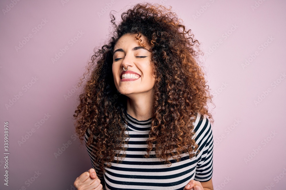 Young beautiful woman with curly hair and piercing wearing casual striped t-shirt very happy and excited doing winner gesture with arms raised, smiling and screaming for success. Celebration concept. Young beautiful woman with curly hair and piercing wearing casual striped t-shirt very happy and excited doing winner gesture with arms raised, smiling and screaming for success. Celebration concept.