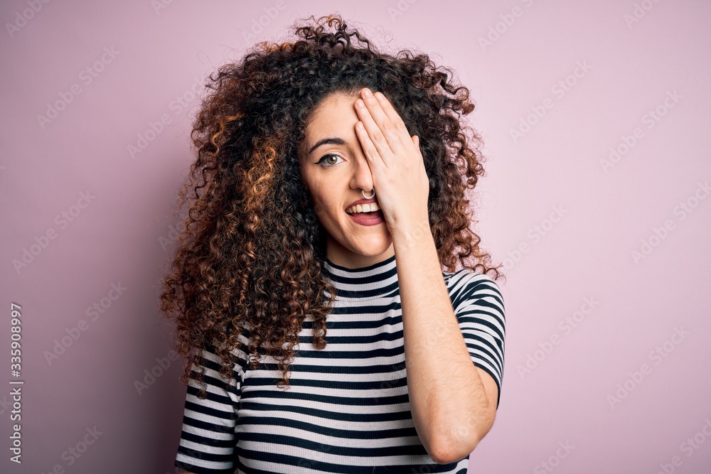 Young beautiful woman with curly hair and piercing wearing casual striped t-shirt covering one eye with hand, confident smile on face and surprise emotion.