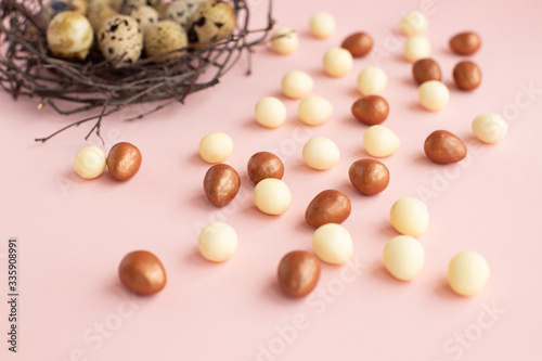 Nest of twigs with quail eggs on light pink background with small eggs made of white, dark chocolate. Easter greeting card, symbol of celebration Sunday of Christ. Holiday composition,congratulations