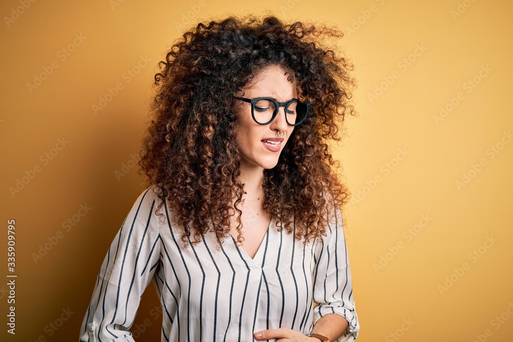 Young beautiful woman with curly hair and piercing wearing striped shirt and glasses with hand on stomach because indigestion, painful illness feeling unwell. Ache concept. Young beautiful woman with curly hair and piercing wearing striped shirt and glasses with hand on stomach because indigestion, painful illness feeling unwell. Ache concept.