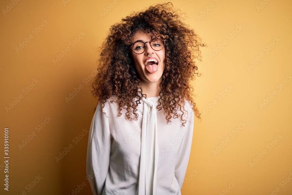 Young beautiful brunette woman with curly hair and piercing wearing shirt and glasses sticking tongue out happy with funny expression. Emotion concept. Young beautiful brunette woman with curly hair and piercing wearing shirt and glasses sticking tongue out happy with funny expression. Emotion concept.