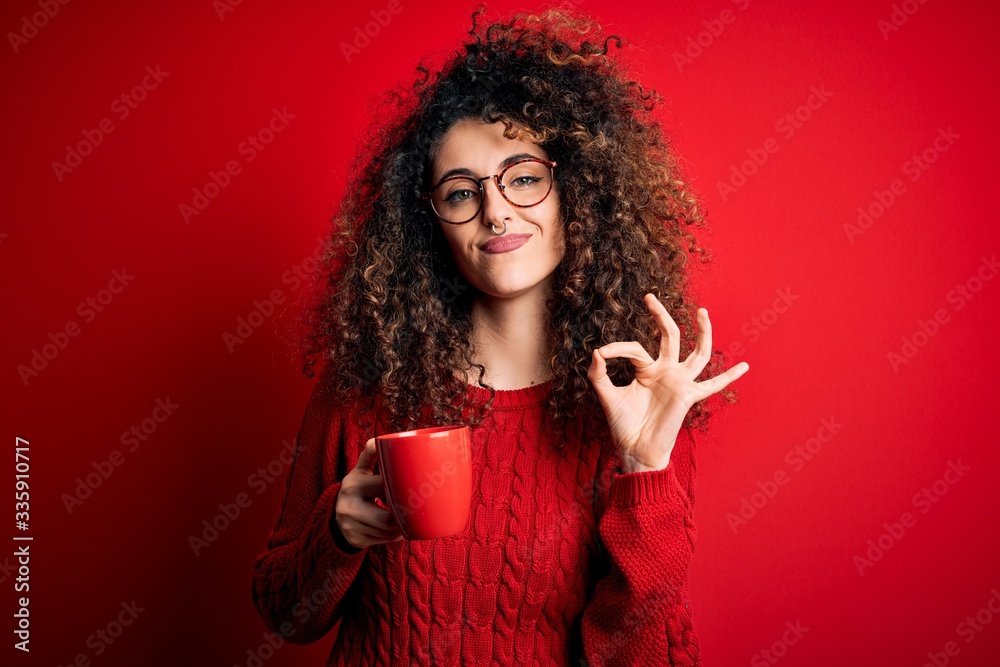 Young beautiful woman with curly hair and piercing drinking red cup of coffee doing ok sign with fingers, excellent symbol Young beautiful woman with curly hair and piercing drinking red cup of coffee doing ok sign with fingers, excellent symbol
