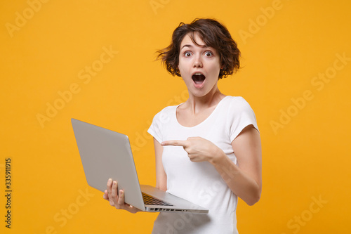 Shocked young brunette woman girl in white t-shirt posing isolated on yellow orange background in studio. People lifestyle concept. Mock up copy space. Pointing index finger on laptop pc computer.
