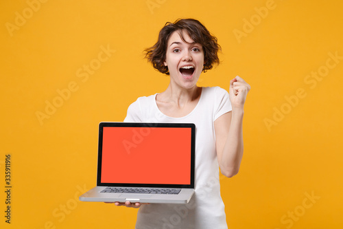 Excited young brunette woman in white t-shirt posing isolated on yellow background. People lifestyle concept. Mock up copy space. Hold laptop pc computer with blank empty screen doing winner gesture.