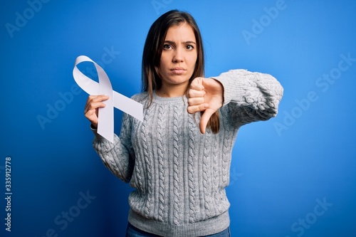 Young blonde woman holding cancer awareness and stop women violence white ribbon with angry face, negative sign showing dislike with thumbs down, rejection concept