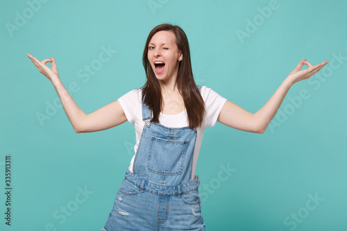 Cheerful young woman girl in casual denim clothes isolated on blue turquoise wall background. People lifestyle concept. Mock up copy space. Hold hands in yoga gesture, relaxing meditating, blinking.