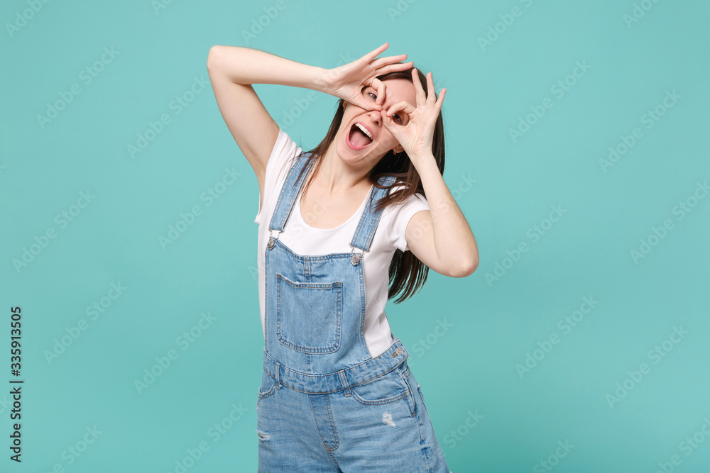 Funny young brunette woman in casual denim clothes posing isolated on blue turquoise background. People lifestyle concept. Mock up copy space. Holding hands near eyes, imitating glasses or binoculars.