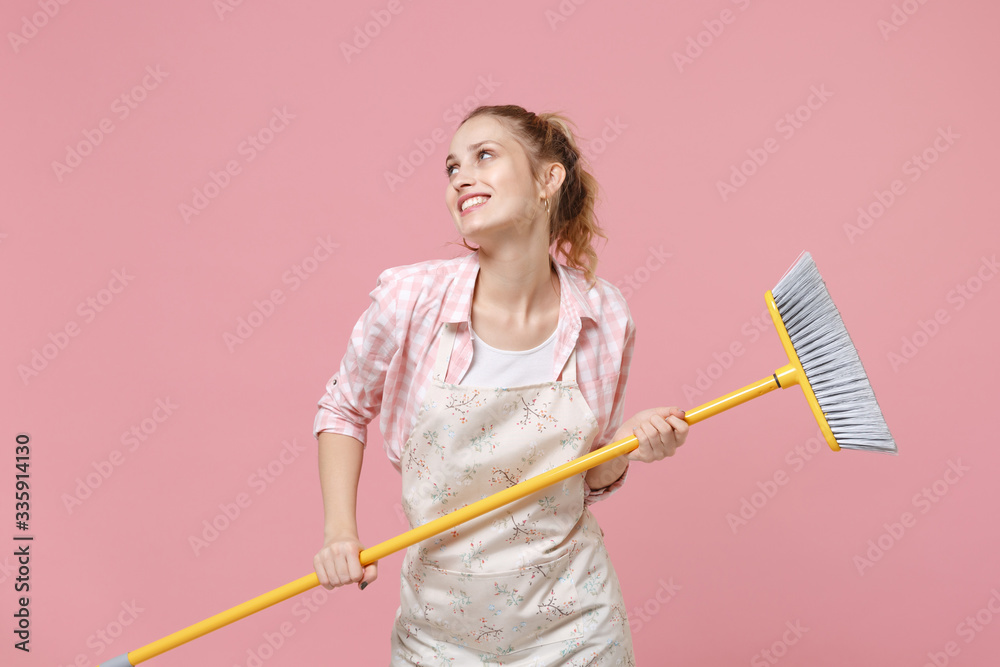 Smiling young woman housewife in casual clothes, apron doing housework isolated on pastel pink background studio portrait. Housekeeping concept. Mock up copy space. Hold in hands broom, looking aside. Smiling young woman housewife in casual clothes, apron doing housework isolated on pastel pink background studio portrait. Housekeeping concept. Mock up copy space. Hold in hands broom, looking aside.