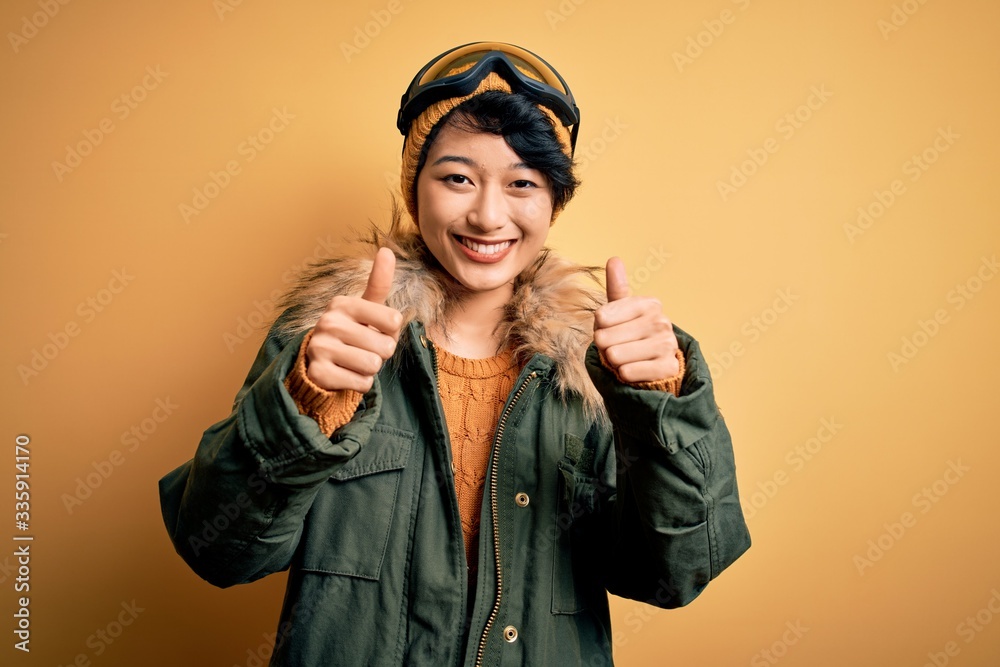 Beautiful asian skier girl wearing snow sportswear using ski goggles over yellow background approving doing positive gesture with hand, thumbs up smiling and happy for success. Winner gesture.