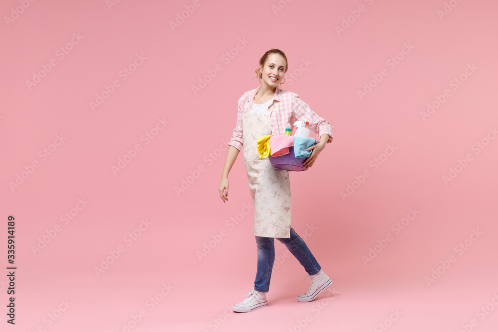 Side view of smiling young woman housewife in apron hold basin with detergent bottles washing cleansers doing housework isolated on pink background in studio. Housekeeping concept. Looking camera. Side view of smiling young woman housewife in apron hold basin with detergent bottles washing cleansers doing housework isolated on pink background in studio. Housekeeping concept. Looking camera.
