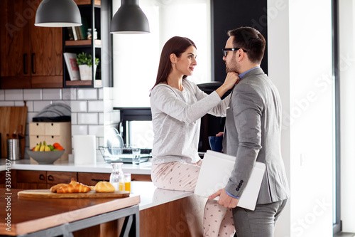 Young modern couple in the kitchen before he gone to work she in pyjama sitting on table and saying goodbye