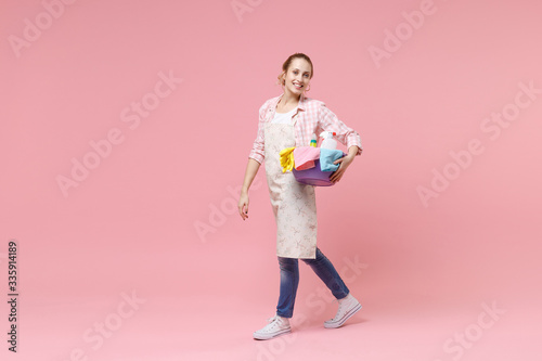 Side view of smiling young woman housewife in apron hold basin with detergent bottles washing cleansers doing housework isolated on pink background in studio. Housekeeping concept. Looking camera.