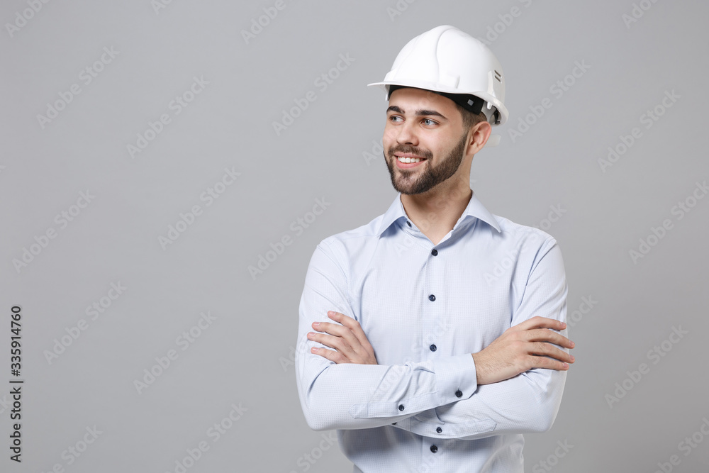 Smiling young unshaven business man in light shirt protective construction helmet isolated on grey wall background. Achievement career wealth business concept. Holding hands crossed, looking aside. Smiling young unshaven business man in light shirt protective construction helmet isolated on grey wall background. Achievement career wealth business concept. Holding hands crossed, looking aside.