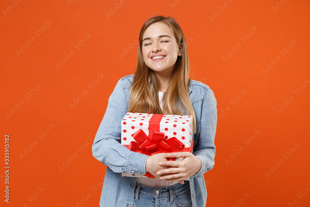 Smiling pensive young woman in casual denim clothes isolated on orange background. Valentine's Day Women's Day birthday holiday concept. Hold red present box with gift ribbon bow, keeping eyes closed. Smiling pensive young woman in casual denim clothes isolated on orange background. Valentine's Day Women's Day birthday holiday concept. Hold red present box with gift ribbon bow, keeping eyes closed.