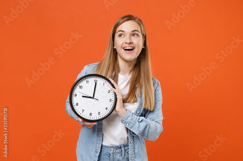 Cheerful young woman girl in casual denim clothes isolated on bright orange wall background studio portrait. People sincere emotions lifestyle concept. Mock up copy space. Hold clock looking aside up.