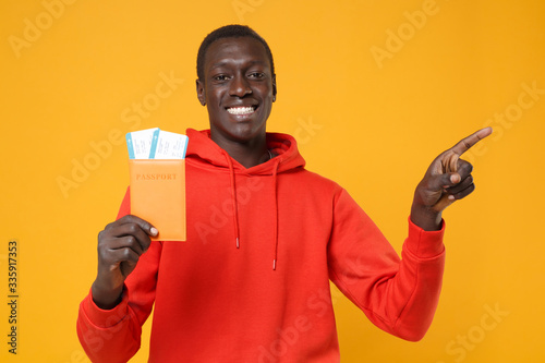 Smiling young african american man in red streetwear hoodie isolated on yellow background. People lifestyle concept. Mock up copy space. Hold passport tickets boarding pass, point index finger aside.