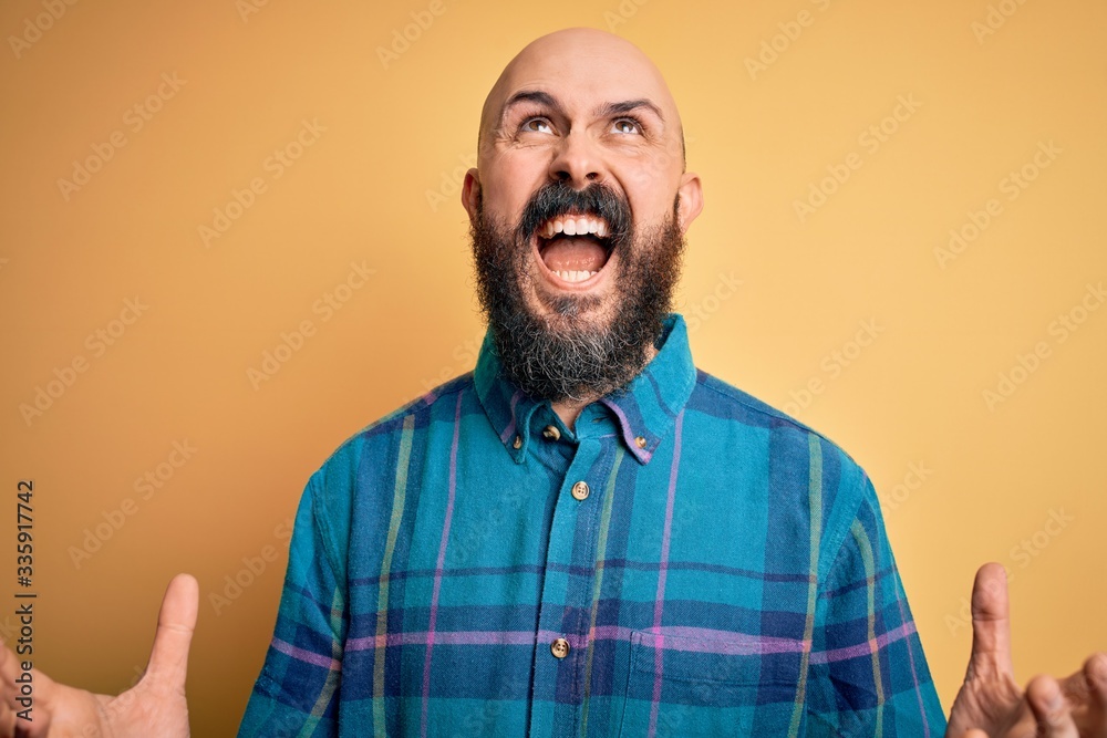 Fototapeta premium Handsome bald man with beard wearing casual shirt standing over isolated yellow background crazy and mad shouting and yelling with aggressive expression and arms raised. Frustration concept.