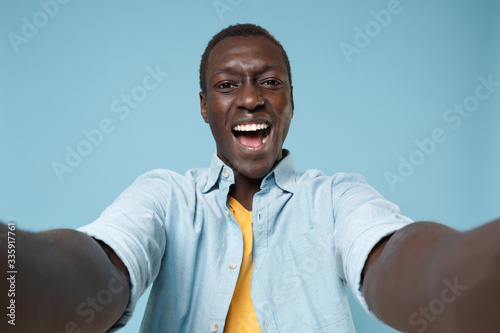 Close up of cheerful young african american man guy in casual shirt, yellow t-shirt posing isolated on blue background. People lifestyle concept. Mock up copy space. Doing selfie shot on mobile phone. © ViDi Studio