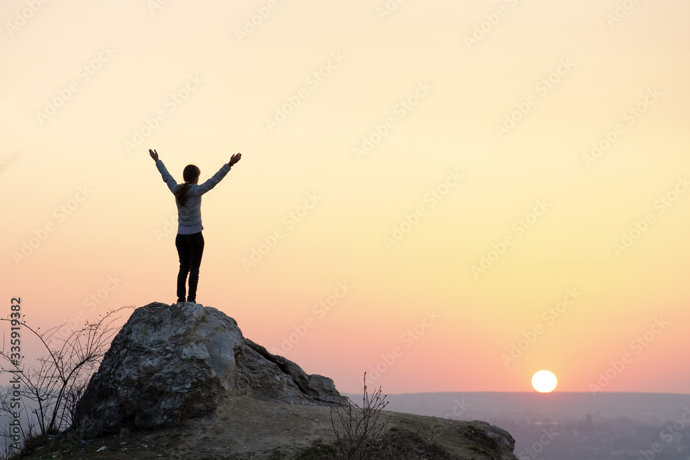 Silhouette of a woman hiker standing alone on big stone at sunset in mountains. Female tourist raising her hands up on high rock in evening nature. Tourism, traveling and healthy lifestyle concept.