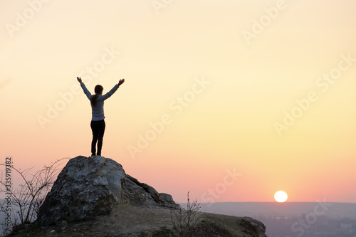 Silhouette of a woman hiker standing alone on big stone at sunset in mountains. Female tourist raising her hands up on high rock in evening nature. Tourism, traveling and healthy lifestyle concept. © bilanol