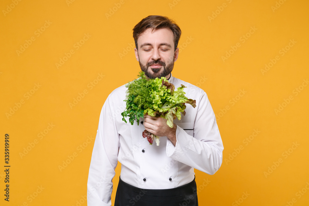 Smiling young bearded male chef cook or baker man in white uniform shirt isolated on yellow wall background. Cooking food concept. Mock up copy space. Hold sniffing bunch of greens salad dill parsley. Smiling young bearded male chef cook or baker man in white uniform shirt isolated on yellow wall background. Cooking food concept. Mock up copy space. Hold sniffing bunch of greens salad dill parsley.
